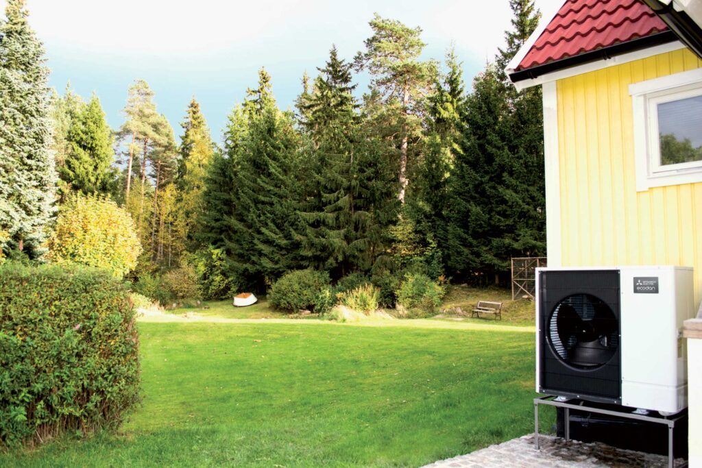 Omakotitalon kulmassa näkyy ilmalämpöpumpun ulkoyksikkö. Taustalla vihreä nurmikko pihamaalla.