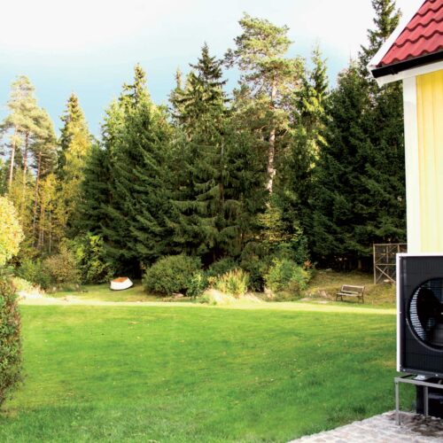 Omakotitalon kulmassa näkyy ilmalämpöpumpun ulkoyksikkö. Taustalla vihreä nurmikko pihamaalla.
